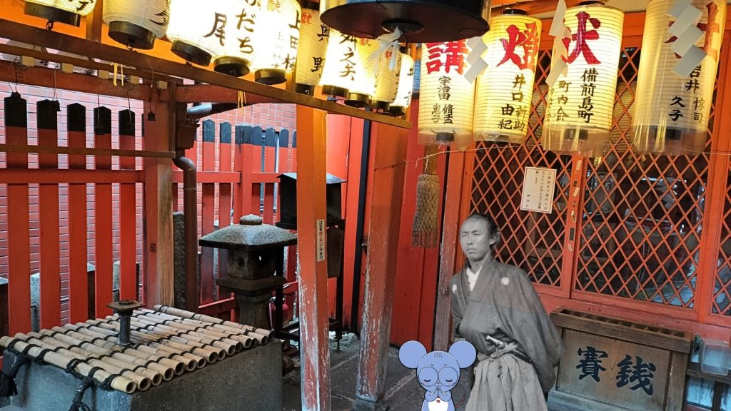京都の土佐藩ゆかりの神社「岬神社（通称：土佐稲荷）」の境内を撮影した写真。赤い鳥居や名前の入った提灯が並ぶ幻想的な雰囲気の中、お賽銭箱の前に坂本龍馬の肖像写真と、お祈りのポーズをしたネズミのキャラクター「ちゅう乃」が並んでいる。中岡慎太郎ランドの京都史跡巡り解説用。