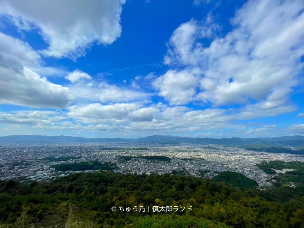 京都・大文字山の火床付近から見渡す京都市街の大パノラマ。青空と山並みの下に町並みが広がる。